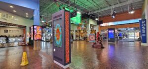 A wide shopping mall concourse featuring multiple tall, black digital kiosks displaying colorful graphics. The brightly lit space includes storefronts like a bakery and a salon under a high, green industrial ceiling.